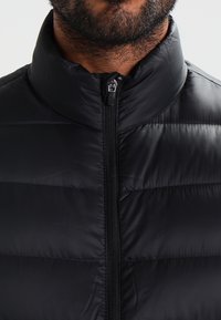 Giacca a vento nera con colletto alto, dotata di chiusura con zip e cuciture orizzontali, realizzata in un materiale sintetico liscio e leggero.