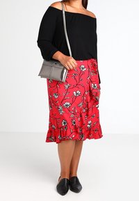 Žena v černém topu bez ramínek, červené květinové midi sukni, černých nazouvacích balerínách a stříbrné crossbody kabelce.