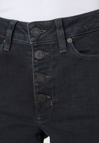 Mörka denimjeans med hög midja, fem stora svarta knappar, synlig söm och en dragkedjedetalj vid gylfen.
