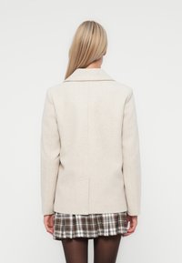 Manteau ajusté beige clair avec col cranté, coupe structurée et texture lisse. La vue arrière montre des lignes épurées et un design minimaliste.