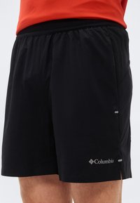 Μαύρο σορτς τρεξίματος Columbia που φοριέται από άτομο με πορτοκαλί μπλούζα, με ελαστική ζώνη στη μέση και μικρές πλευρικές σχισμές.