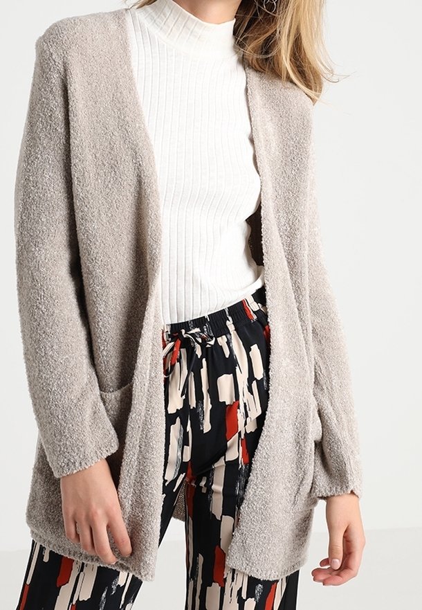 Femme portant un cardigan ouvert beige texturé, un col roulé blanc côtelé et un pantalon à imprimé abstrait noir, rouge et beige.