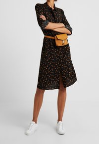 Czarna sukienka z pomarańczowym wzorem w klucze, zapinana na guziki, z podwiniętymi rękawami i rozporkiem z przodu. Zestawiona z beżową torebką typu crossbody i białymi sneakerami.