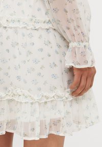 Lättviktig vit klänning med blå blommönster, volangkant och volanger på ärmarna, tillverkad av genomskinligt tyg. Har elastiska ärmslut och en rynkad midja.