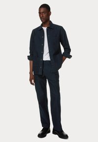 Donkerblauwe op maat gemaakte blazer met knoopsluiting en zak, gecombineerd met bijpassende op maat gemaakte broek. Witte t-shirt met ronde hals eronder gedragen.