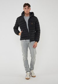 Giacca a vento nera con zip frontale, cappuccio staccabile e polsini a costine. Presenta una tasca sul petto con dettaglio del logo. Camicia grigia e jeans sotto.