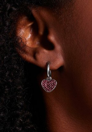 Close-up van een oor met een donkere huid en kort krullend haar, met een zilveren oorring met een fonkelende roze hartvormige hanger.