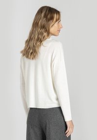 Kobieta z falującymi blond włosami, ubrana w biały sweter z długim rękawem i szare spodnie, stojąca bokiem na jasnym, jednolitym tle.