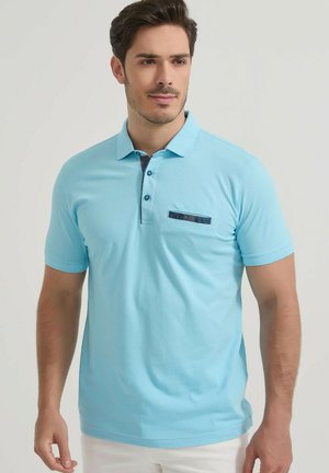Homme portant un polo bleu clair à manches courtes avec une patte de boutons sombre et un détail horizontal sombre sur la poche poitrine.