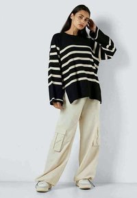 Γυναίκα που φοράει μια υπερμεγέθη μαύρη ζακέτα με φαρδιές λευκές ρίγες και χαλαρές μπεζ cargo παντελόνες με λευκά αθλητικά παπούτσια, που στέκεται μπροστά σε έναν απλό τοίχο.