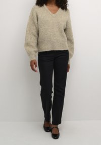 Světlý béžový pletený svetr s výstřihem do V a nadýchanými rukávy, kombinovaný s černými kalhotami se vzpřímenými nohavicemi a černými botami na podpatku.