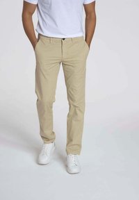 Chinos beige avec une coupe ajustée, en tissu coton, dotés d'une fermeture à bouton et de poches standard, associés à des baskets blanches.