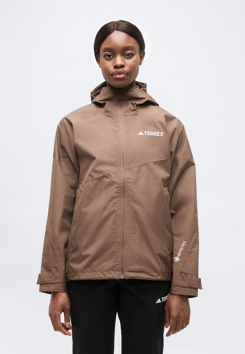 Femme portant une veste Terrex GORE-TEX marron à capuche, fermeture éclair à l'avant, et un pantalon Terrex noir, sur un fond gris uni.