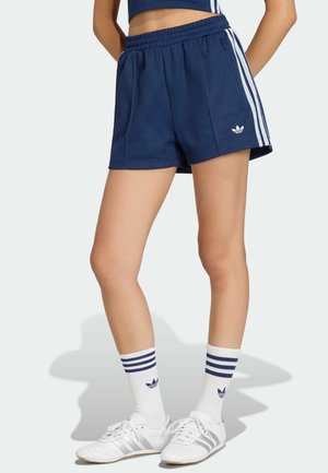 Osoba ubrana w granatowe szorty Adidas z białymi paskami, białe skarpetki Adidas z niebieskimi paskami oraz białe buty sportowe Adidas.