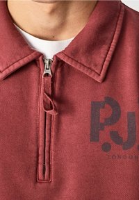 Červená mikina na zip s límečkem, s logem ve tmavém potisku. Vyrobena z texturované látky, ukazuje švy a kovové detaily zipu.
