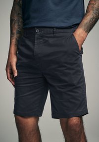 Námořnické modré kraťasy vyrobené z měkké tkaniny, s knoflíkovým zapínáním, bočními kapsami a volným střihem, končícími těsně nad kolenem.