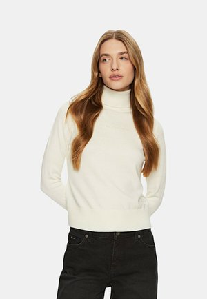 Maglione bianco a collo alto con polsini e fondo a coste, con logo in rilievo sul davanti, abbinato a jeans neri. Tessuto liscio e morbido.