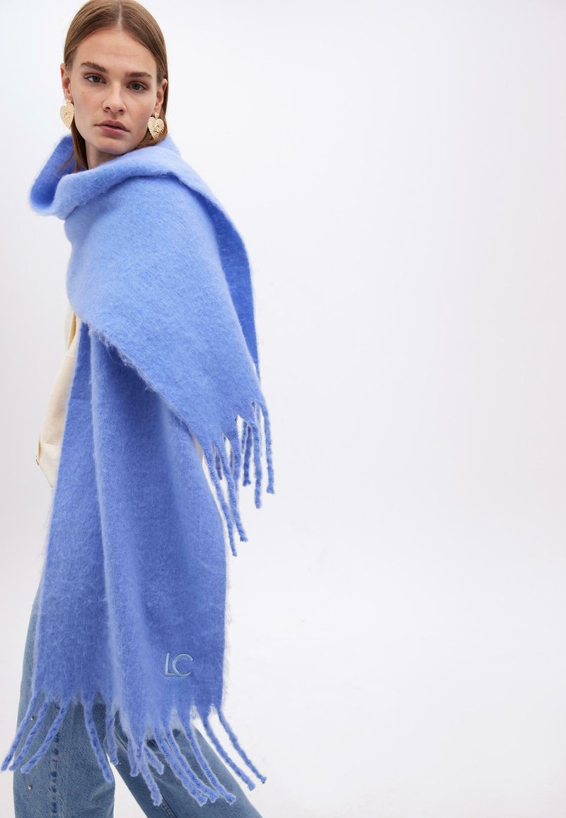 Lyseblå uldne sjal med frynset kanter og broderet logo. Blød tekstur og draperet design, båret over en cremefarvet top og jeans.
