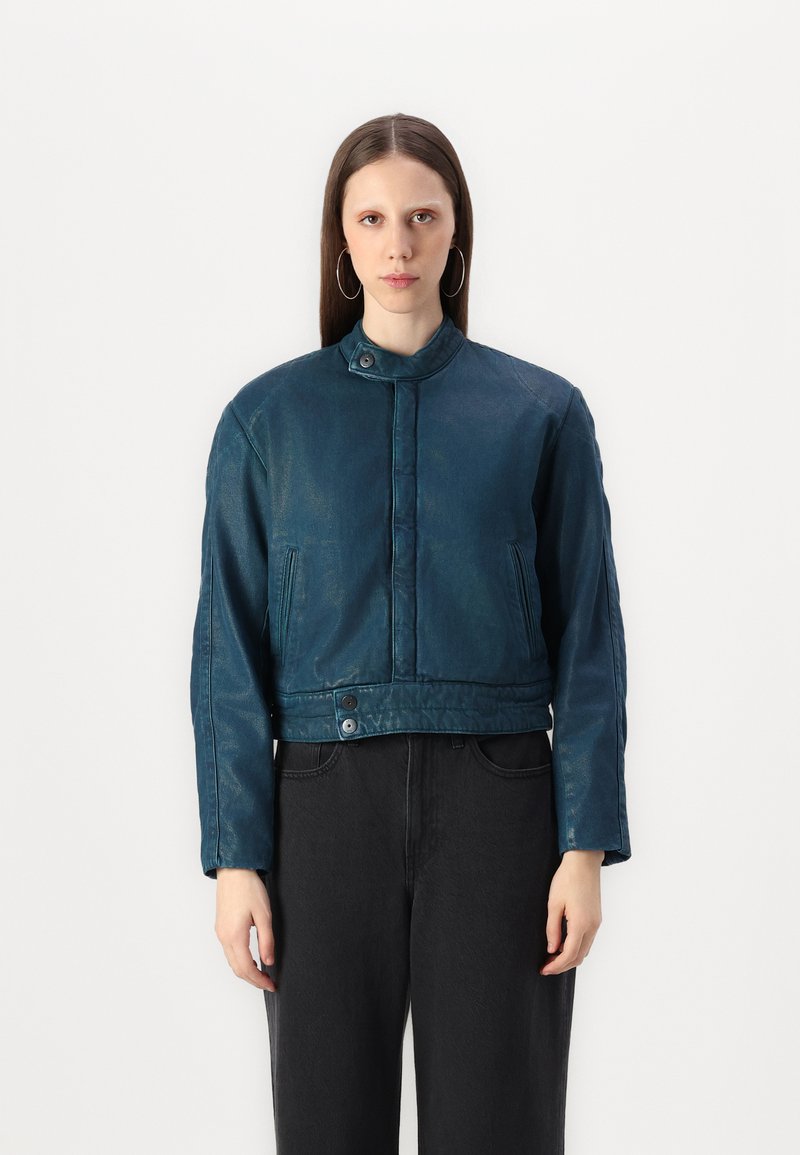 Chaqueta de cuero verde azulado recortada con un alto cuello, cierre de botones y bolsillos laterales; presenta una textura suave y un diseño minimalista.