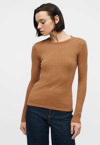 Γυναίκα με κοντά σκούρα μαλλιά που φοράει εφαρμοστή καφέ μακρυμάνικη μπλούζα με ρίγες και σκουρό μπλε τζιν, κοιτάζοντας προς τα κάτω και ελαφρώς προς το πλάι.