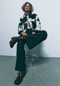 Veste en fausse fourrure à motif vache, noire et blanche ; large pantalon noir ; bottines marron brillantes ; assise sur un tabouret chromé devant un fond clair.
