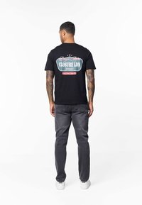 Svart bomulls-T-shirt med grafisk design på baksidan, med texten "CLOSURE LDN" i vitt och rött, kombinerad med mörkgrå jeans och vita sneakers.