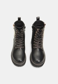 Botas de tobillo de cuero negro con herrajes en tono dorado, que presentan un cierre de cordones frontal y cremallera lateral. Cordones texturizados y cuello acolchado. Suela de goma.