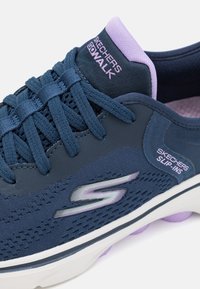 Marinefarbene Skechers Go Walk Slippers mit strukturiertem Mesh-Obermaterial, lila Akzenten, Schnürsenkeln und einer gepolsterten Sohle mit runden Details.