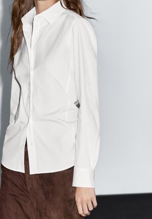 Personne portant une chemise blanche boutonnée et un pantalon brun foncé sur un fond clair uni, cadrée du cou à la mi-cuisse.