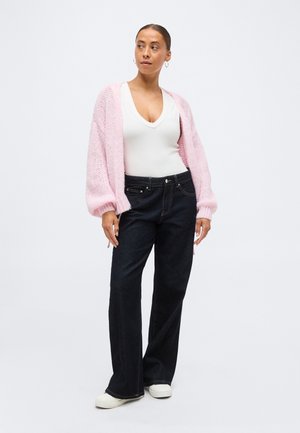 Femme debout portant un haut blanc à col en V profond, un cardigan rose en maille épaisse, un jean noir à jambes larges, des baskets blanches et des boucles d'oreilles créoles.