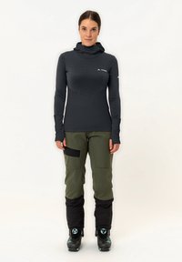 Schwarzes Langarmoberteil mit gerippter Textur und Kapuze, kombiniert mit grünen Hosen mit schwarzen Akzenten und wasserdichten Stiefeln.