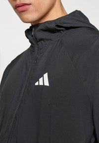 Zwarte jas met capuchon, voorzien van een ritssluiting aan de voorkant, een gladde textuur en een wit logo met drie strepen op de linkerborst.