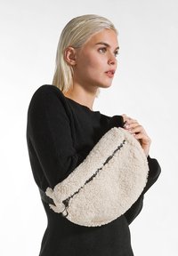 Borsa in finta pelliccia color crema con forma curva e zip nera. Dotata di una tracolla rimovibile per diverse opzioni di trasporto.
