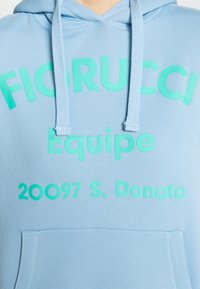 Sudadera con capucha azul claro con un bolsillo delantero, con texto en diversas tonalidades de verde: "FIORUCCI Equipe 20097 S. Donato." Textura de tela suave.