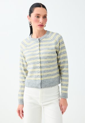 Vrouw met donker haar draagt een grijze cardigan met gele en witte horizontale strepen en witte broek, staand tegen een effen achtergrond.