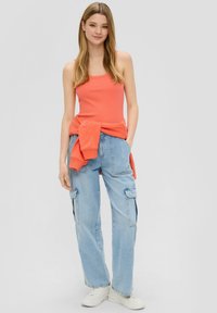 Ein orangefarbenes, geripptes Tanktop, kombiniert mit hellblauen Cargo-Jeans. Der Look wird durch einen um die Taille gebundenen Hoodie und weiße Sneaker ergänzt.