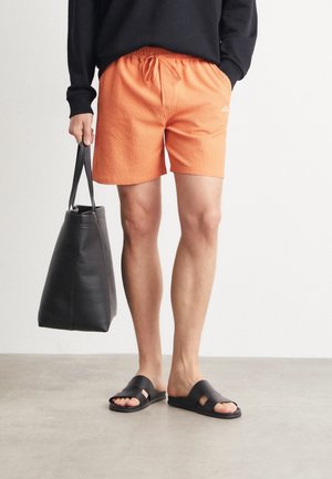 Person iført orange teksturerede shorts, sort sweater, sorte slide-sandaler, bærende en stor sort tote-taske, stående på et glat gulv.