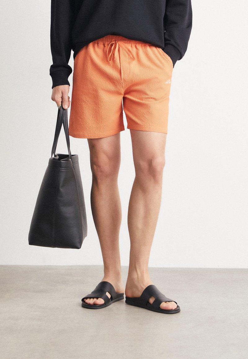 Personne portant un short orange texturé, un pull noir, des sandales noires à enfiler, tenant un grand sac fourre-tout noir, debout sur un sol lisse.