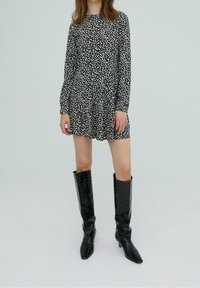 Schwarzes langärmliges Minikleid mit weißem Blumenmuster, geraffter Taille und runder Ausschnitt, kombiniert mit glänzenden schwarzen kniehohen Stiefeln.