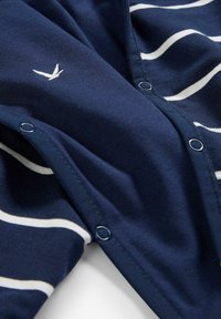 Tessuto blu navy con righe orizzontali bianche, dotato di bottoni a scatto in metallo e un piccolo logo di uccello ricamato. Texture morbida e design casual.