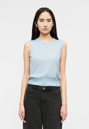 Γυναίκα με μακριά σκούρα μαλλιά, φορόντας αμάνικη ανοιχτό μπλε μπλούζα και μαύρο τζιν ψηλόμεσο παντελόνι, στέκεται με τα χέρια χαλαρά στα πλάγια.