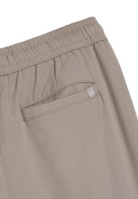 Spodnie z elastycznym pasem w jasnym beżowym materiałem, z tylną kieszenią z małą metką z logo. Gładka tekstura bez widocznych wzorów.