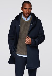 Abrigo de un tono azul marino oscuro con capucha, que presenta un cierre frontal de cremallera y botones. Debajo, un suéter texturizado de color oliva y una camisa de color azul claro con cuello.