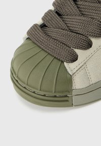 Zaļš un bēšs sporta apavu modelis ar teksturētu gumijas purngali, brūnu pīti šņorēm un mīkstu zamšādas virsmu ar toņiem saskaņotām šuvēm.