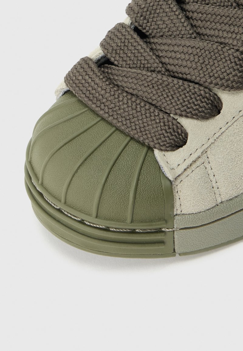 Baskets vertes et beige avec un embout en caoutchouc texturé, des lacets marron tressés et une tige en daim doux avec des coutures ton sur ton.