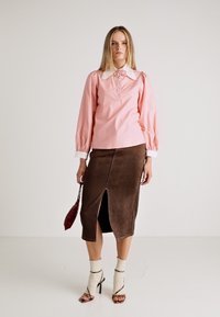 Camicia rosa con maniche ampie e colletto bianco, abbinata a una gonna in suede marrone con uno spacco frontale. Modella che indossa tacchi alti.