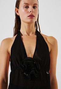 Juoda halter tipo suknelė su raukšlėta tekstūra ir ryškia audinio gėlių detale krūtinėje. Apima elegantišką siuvimą ir dizainą.