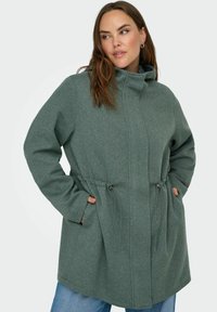 Zelena kapuca iz teksturirane tkanine, s naglašenim vezicama u struku i džepovima sa svake strane. Prikladan, opušten kroj.