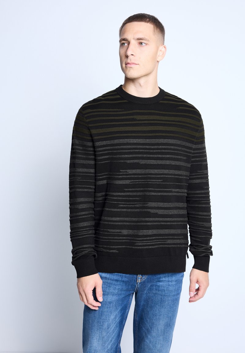 Black sweater with horizontal green and gray stripes, ribbed neckline and cuffs, paired with blue jeans, displayed against a light background.
