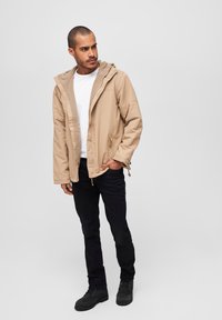Beige hettejakke med glidelås foran, to sidelommer og snortrekkdetaljer, kombinert med en hvit t-skjorte og svarte jeans. Svarte støvler.